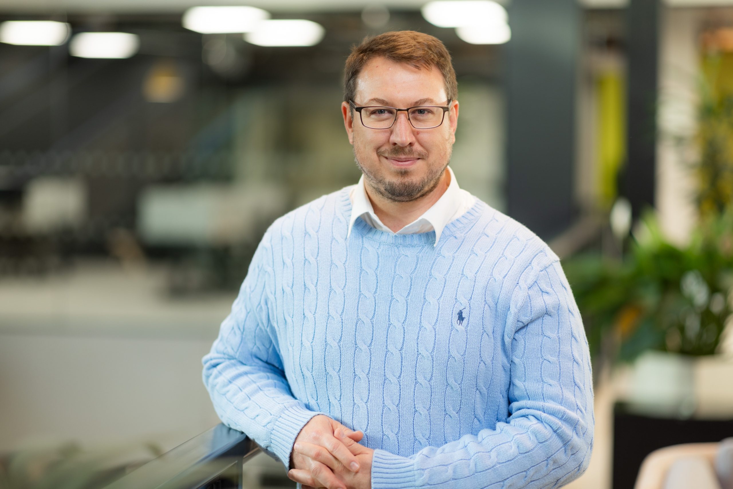 A person standing indoors, wearing a light blue cable-knit sweater over a white collared shirt. The background shows an office environment with blurred details, including desks, chairs, and plants. The individual has their hands clasped in front of them and is positioned slightly to the left of the center of the image.